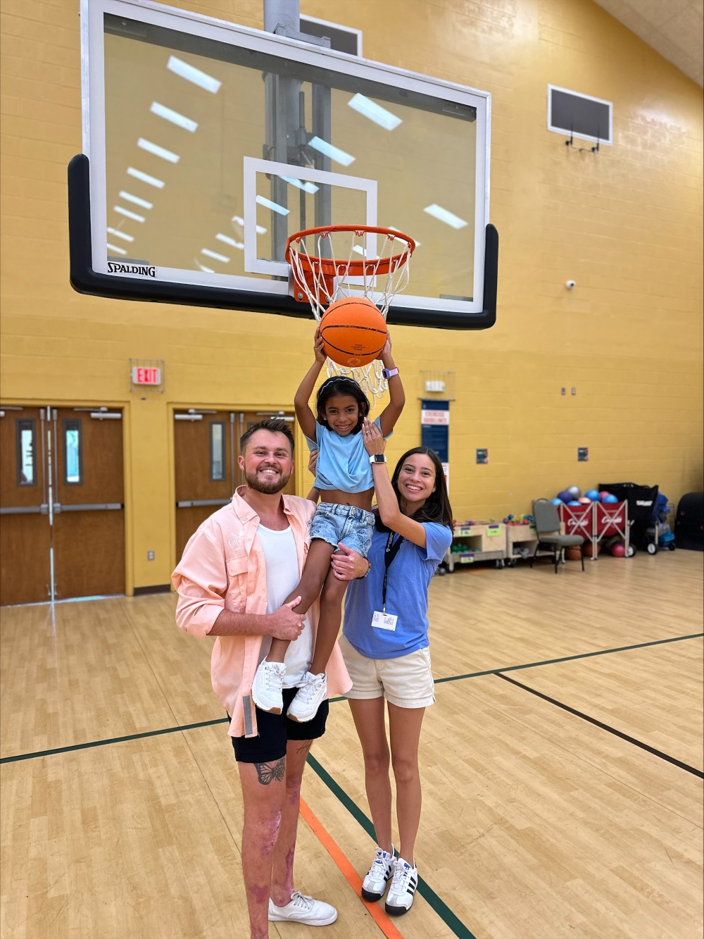 Two adults lift a child to dunk a basketball on an indoor gym hoop